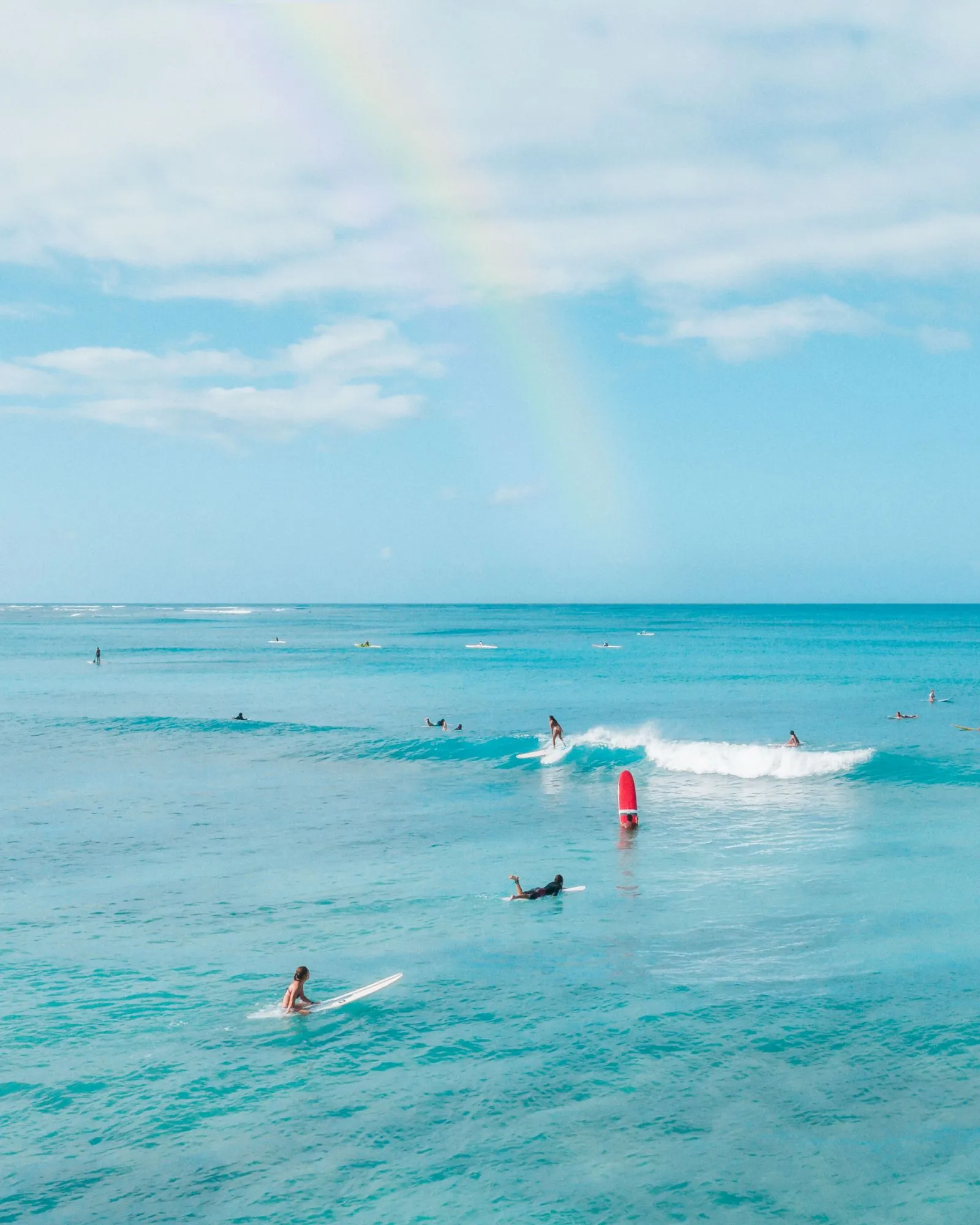 Surfers on longboards in turquoise water, one riding a small wave, with a rainbow overhead.